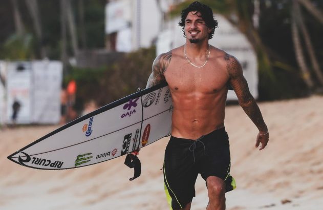 Homem sem camisa e tatuado sorri, caminhando na praia com prancha de surf (logotipos visíveis).