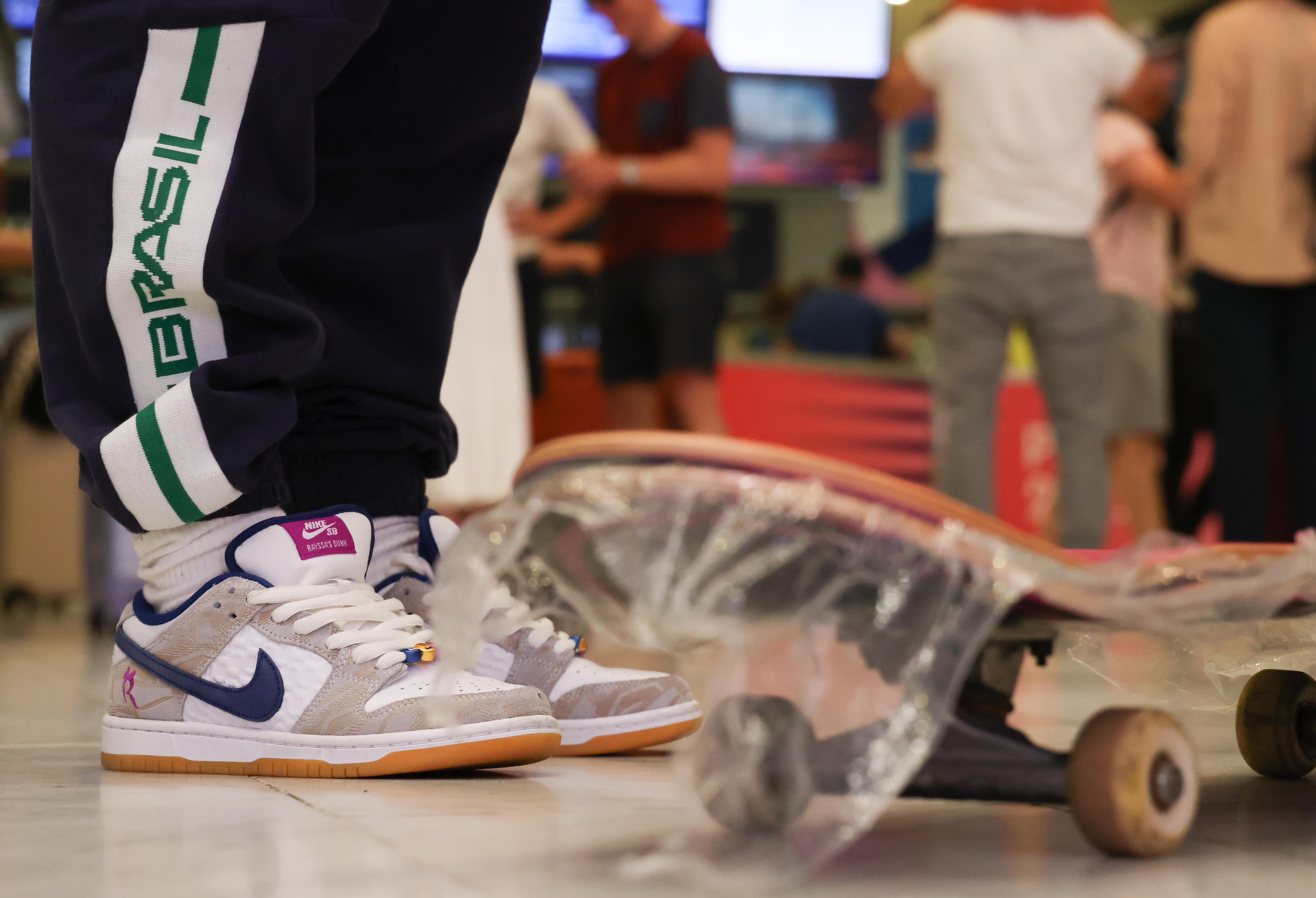 Pessoa de tênis Nike SB e calça 'BRASIL' em pé, com skate embalado no chão.