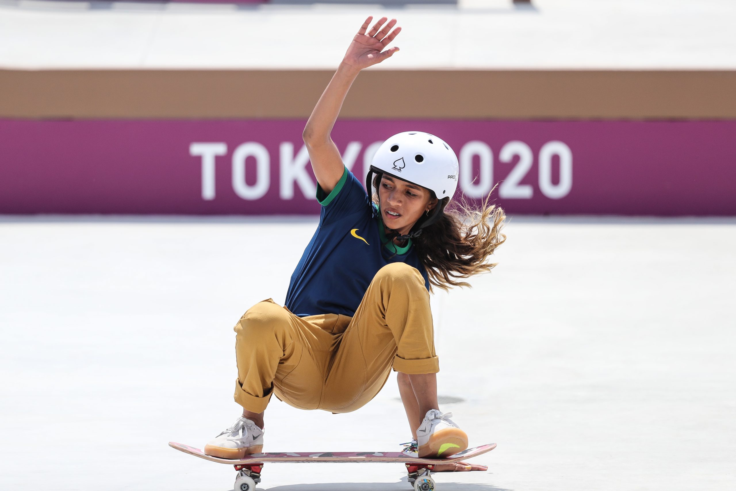 Jovem skatista com capacete agachada no skate, com TOKYO 2020 ao fundo em uma pista.