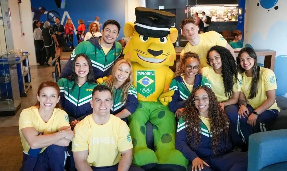 Grupo de atletas do Time Brasil e mascote posam sorrindo em ambiente interno. Vestem uniformes verde, amarelo e azul.