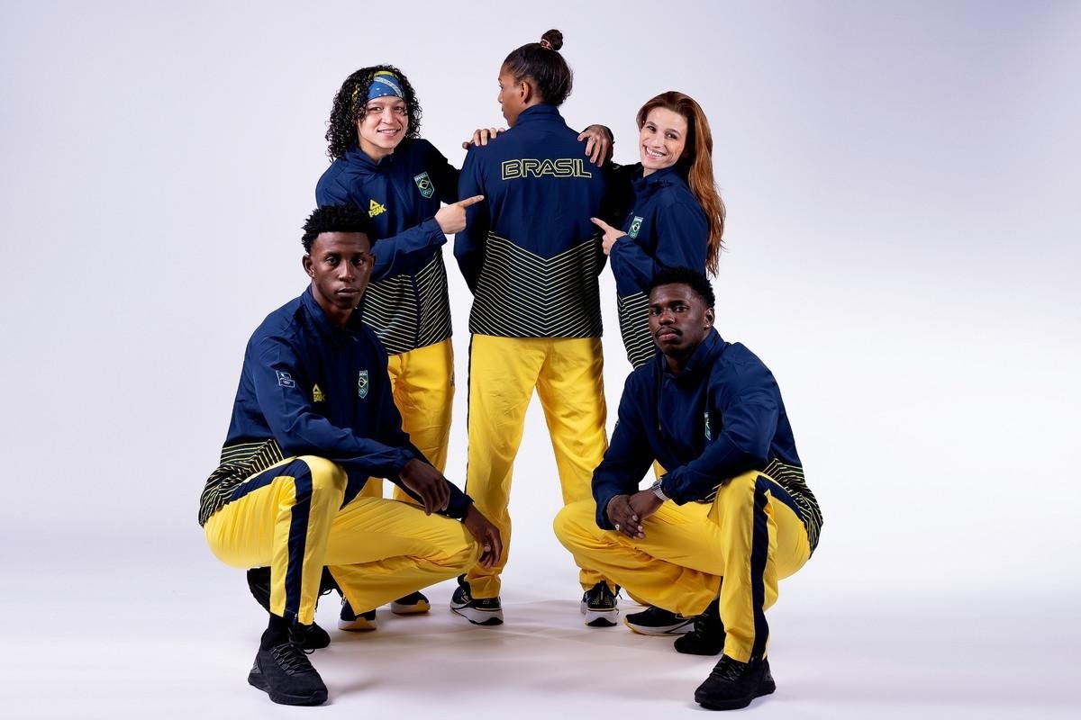 Cinco pessoas em uniformes de treino azuis e amarelos. Três de pé apontam para o agasalho com BRASIL; dois agachados.