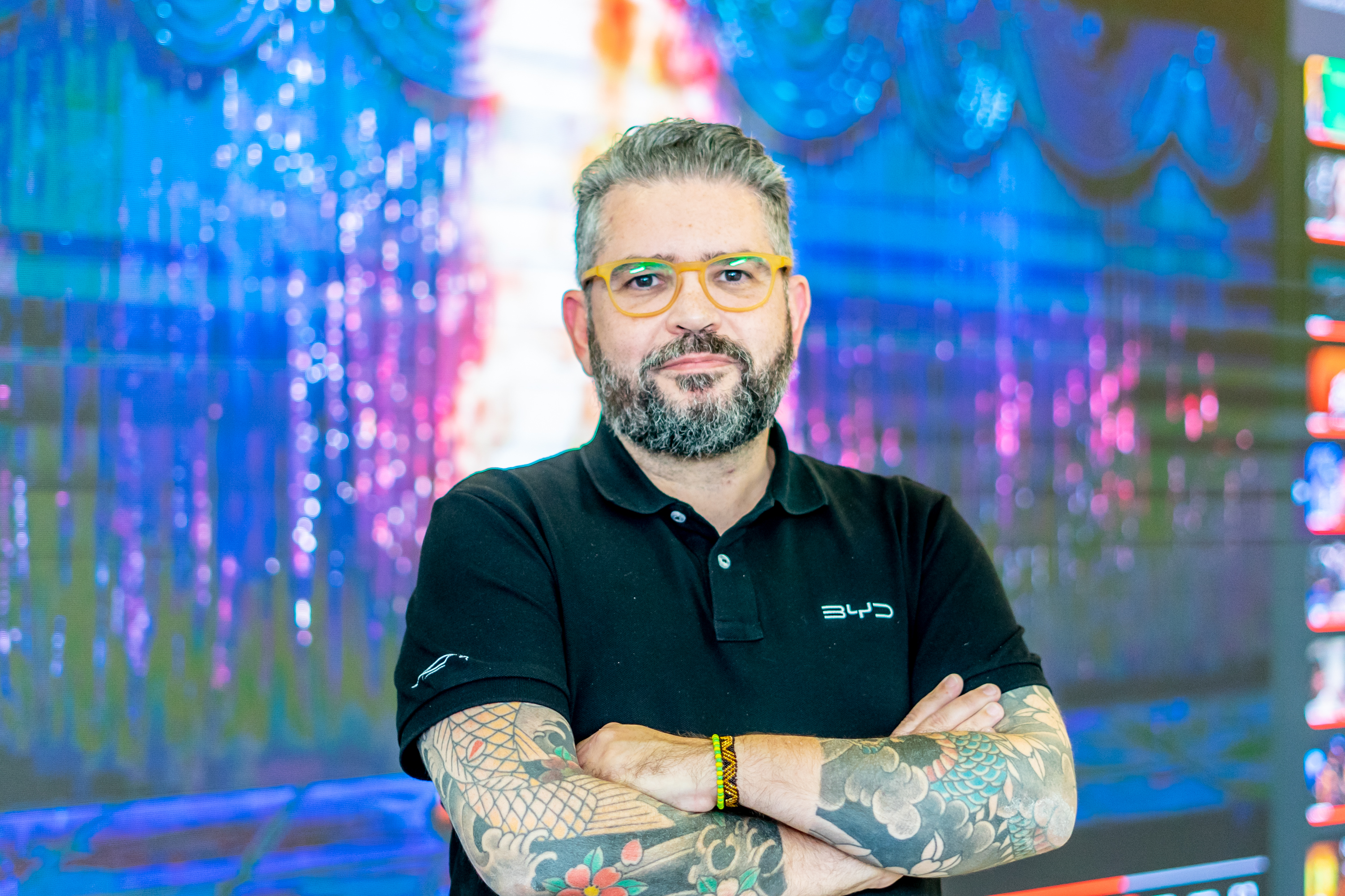Homem barbudo de óculos amarelos, braços cruzados e tatuados. Veste camisa polo preta, fundo com luzes coloridas.