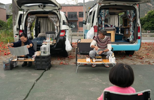 Pessoas ao lado de vans abertas, cozinhando e relaxando ao ar livre. Uma criança observa a cena.
