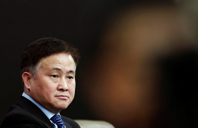 Homem asiático de terno e gravata azul, olhando à esquerda. Fundo escuro e desfocado.