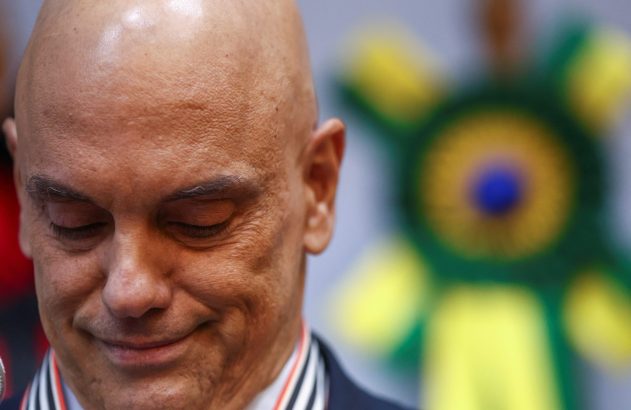 Homem calvo, de olhos baixos, com medalha. Ao fundo, boneco verde e amarelo desfocado.