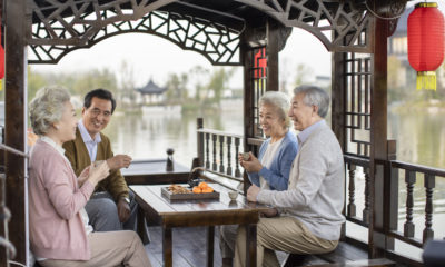 Quatro idosos asiáticos sorriem em barco, tomando chá com lanches. Lago e construção chinesa ao fundo.
