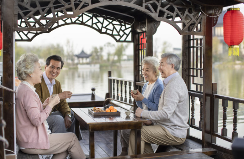 Quatro idosos asiáticos sorriem em barco, tomando chá com lanches. Lago e construção chinesa ao fundo.