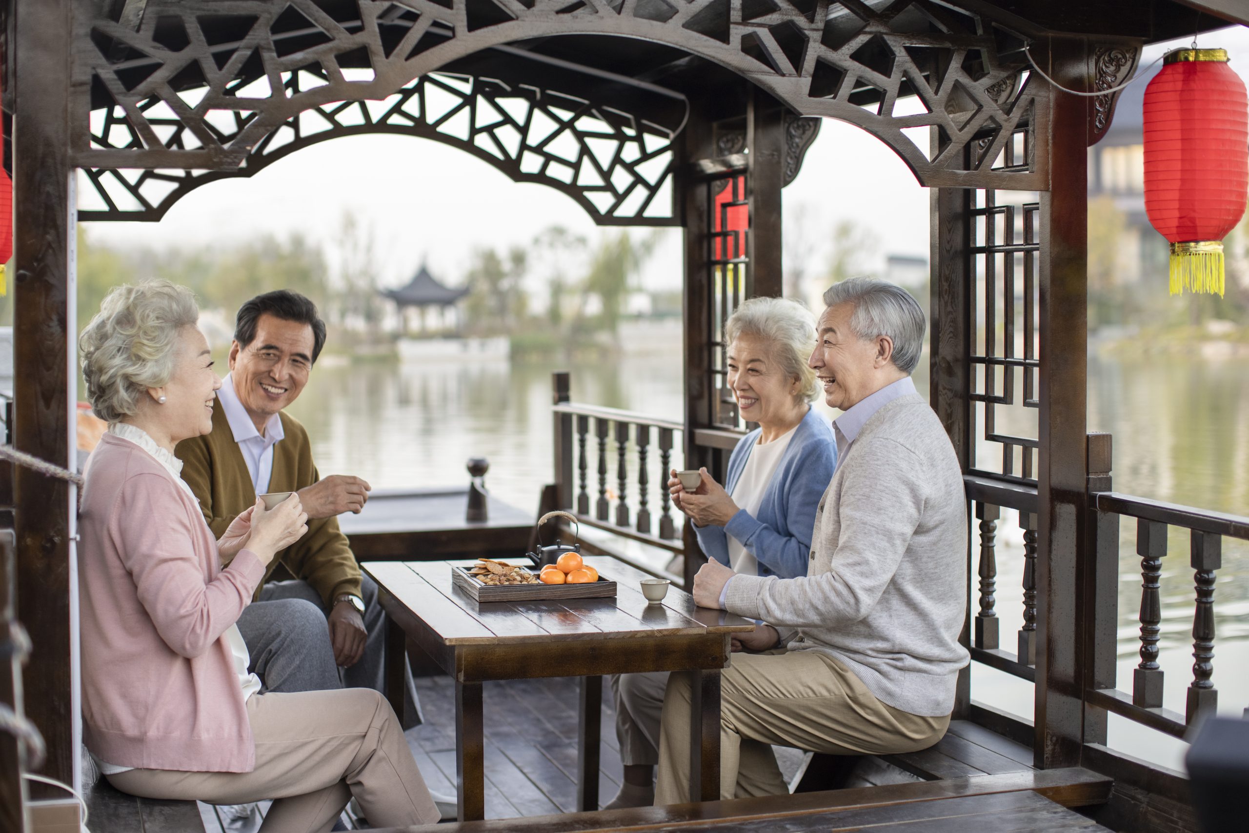 Quatro idosos asiáticos sorriem em barco, tomando chá com lanches. Lago e construção chinesa ao fundo.