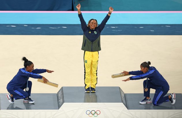 Três atletas no pódio olímpico. A medalhista de ouro do Brasil celebra; outras duas agacham com caixas. Paris 2024.