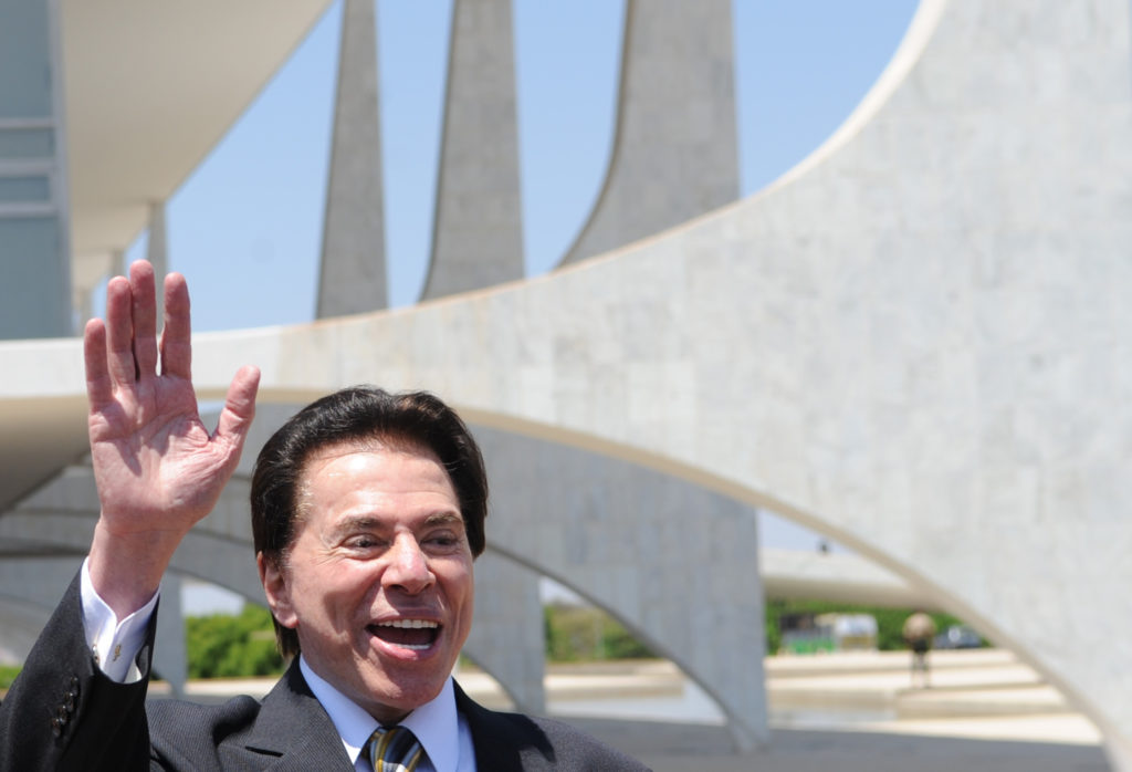 Silvio Santos sorrindo acena em frente a prédio branco moderno.