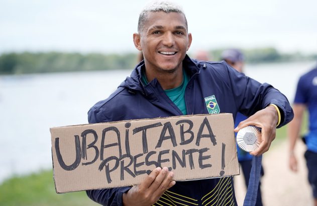 Atleta brasileiro sorrindo, segurando placa "Ubaitaba Presente!" e medalha de prata.