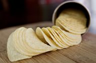 Batatas chips em fila saindo de um pote cilíndrico sobre uma tábua de madeira.