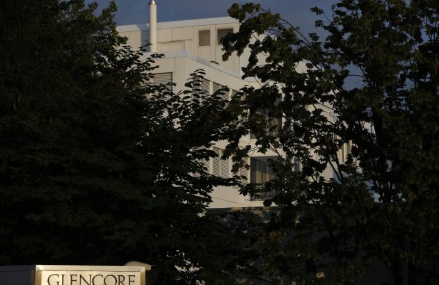 Edifício branco iluminado, parcialmente obscurecido por árvores escuras. Placa 'GLENCORE' visível no canto inferior esquerdo.