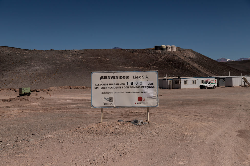 Sinal em campo árido, com construções à direita e uma montanha com estruturas no topo ao fundo.