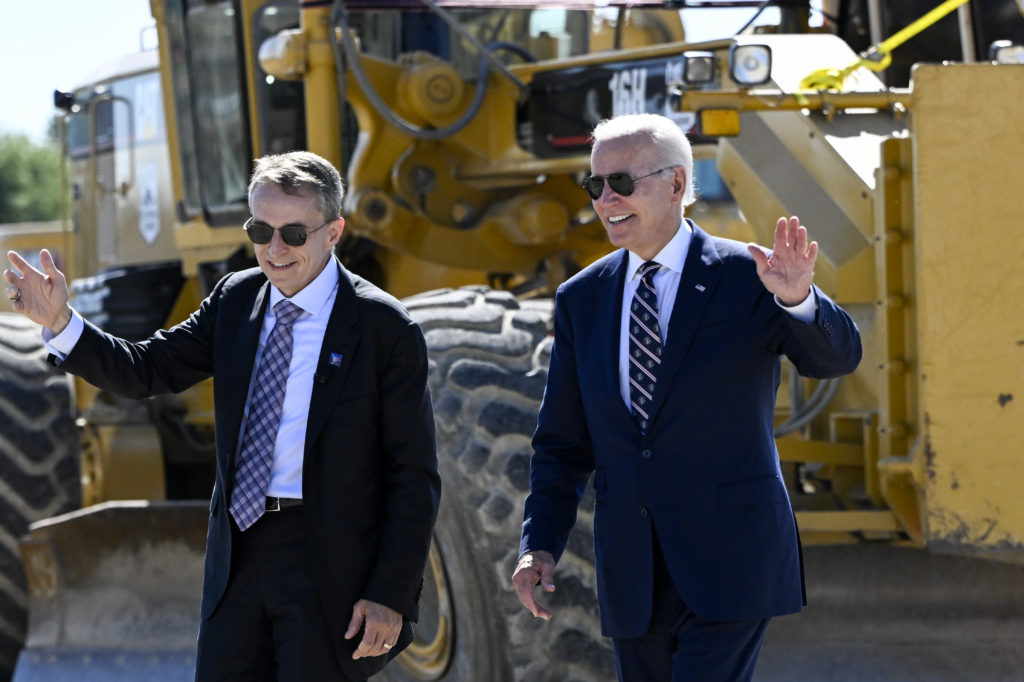 Joe Biden e um CEO acenam sorridentes, caminhando ao lado de equipamento pesado amarelo.