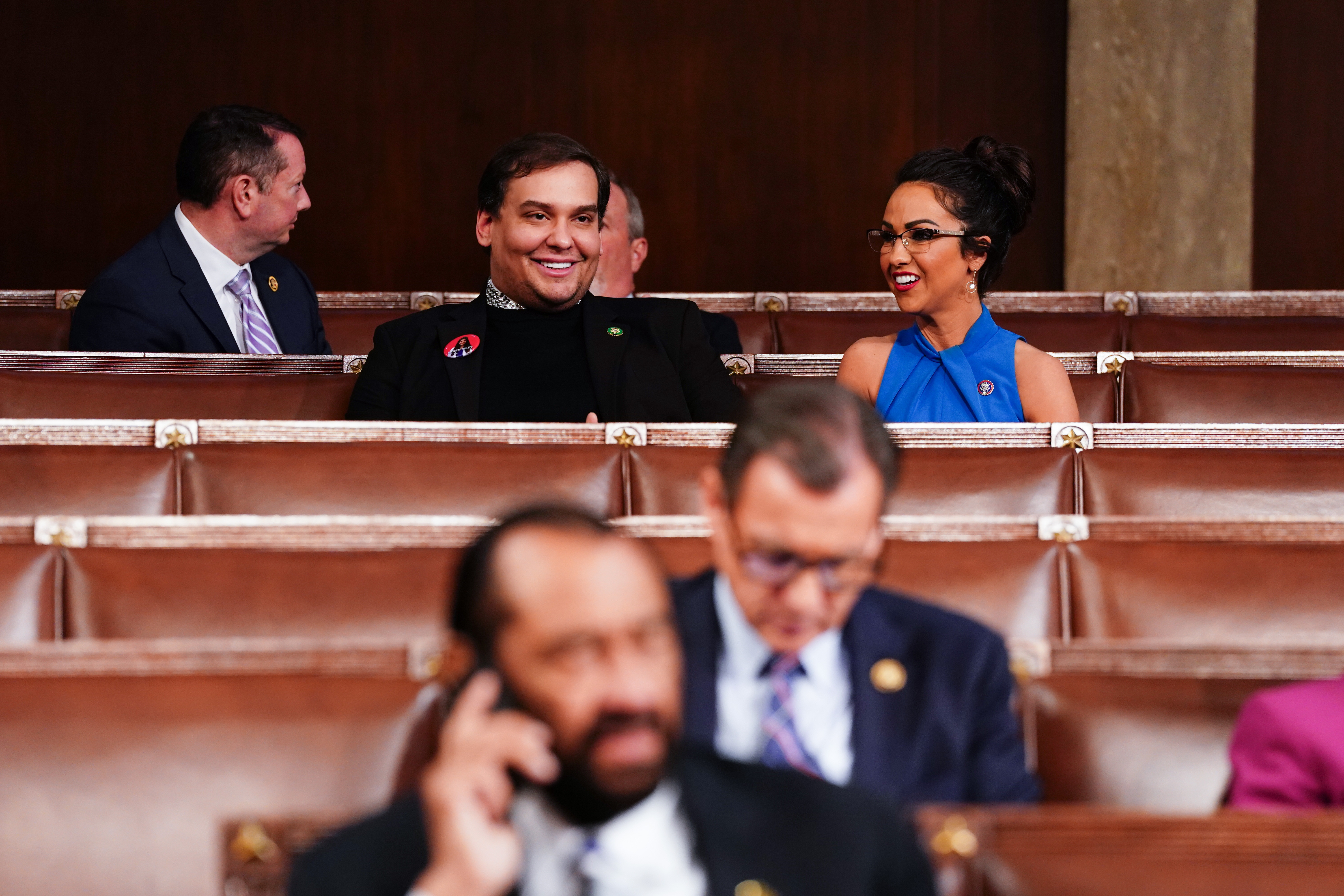 Em um plenário, um homem de preto e uma mulher de azul trocam sorrisos. Outro homem de terno observa.