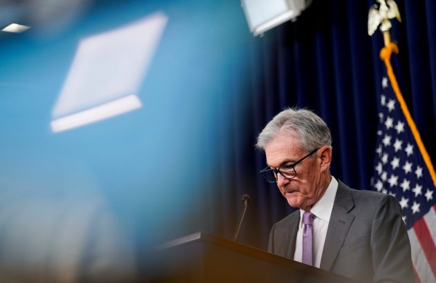 Jerome Powell em pódio, olhando para baixo, com bandeira dos EUA e reflexos de luz em fundo azul.