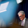 Jerome Powell em pódio, olhando para baixo, com bandeira dos EUA e reflexos de luz em fundo azul.