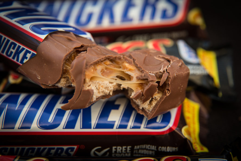 Barra de chocolate Snickers quebrada revelando caramelo e nougat. Outras embalagens Snickers ao fundo.