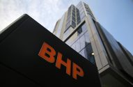 Letreiro preto com 'BHP' laranja e um prédio moderno de vidro em perspectiva de baixo para cima.