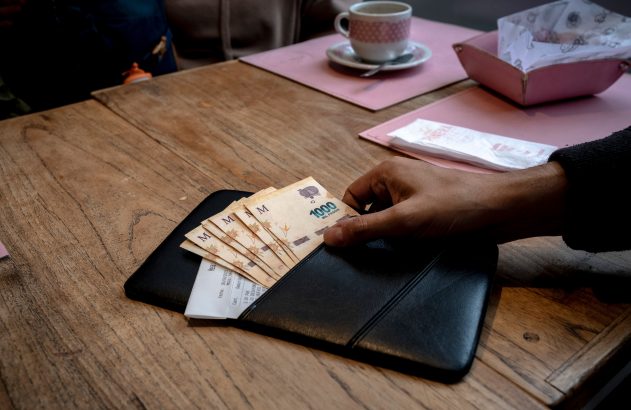 Mão interagindo com notas de dinheiro e recibo em carteira preta sobre mesa de madeira.