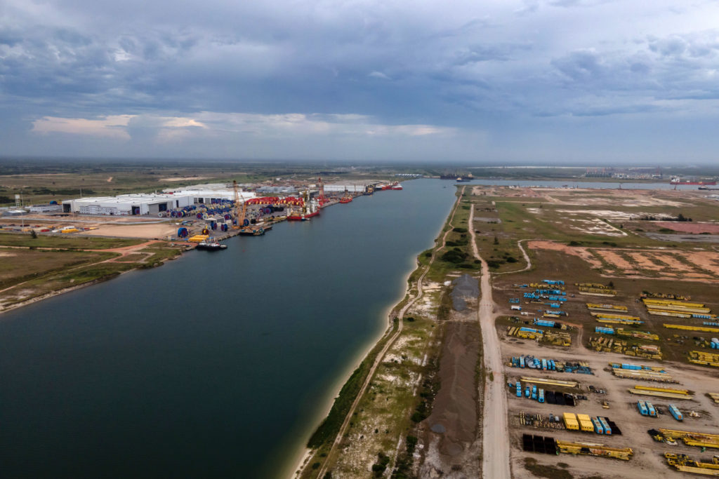 Vista aérea de um porto industrial. Grandes bobinas, navios e guindastes se alinham ao longo de um canal largo.
