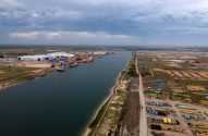 Vista aérea de um porto industrial. Grandes bobinas, navios e guindastes se alinham ao longo de um canal largo.