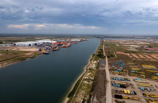 Vista aérea de um porto industrial. Grandes bobinas, navios e guindastes se alinham ao longo de um canal largo.