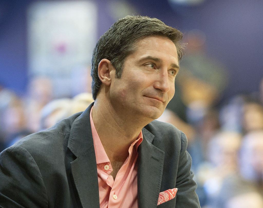 Homem com blazer e camisa rosa olhando pensativamente para o lado em um evento.