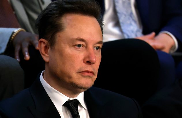 Elon Musk, de terno escuro e gravata preta, sentado e com olhar sério.