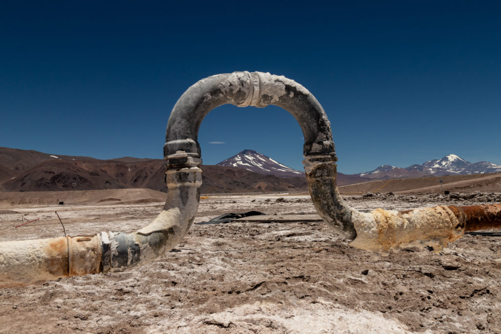 Cano industrial corroído em forma de U em salar árido com montanhas ao fundo e céu azul.