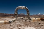 Cano industrial corroído em forma de U em salar árido com montanhas ao fundo e céu azul.