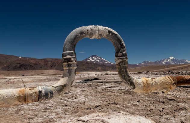 Cano industrial corroído em forma de U em salar árido com montanhas ao fundo e céu azul.
