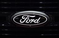 Emblema oval da Ford, preto com letras e bordas brancas, fixado na grade preta de um veículo, com câmera frontal.