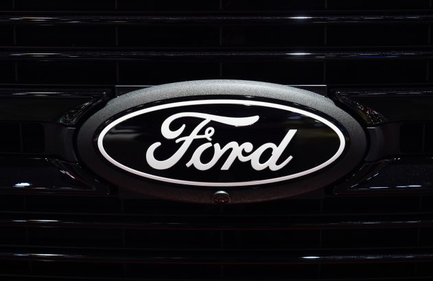 Emblema oval da Ford, preto com letras e bordas brancas, fixado na grade preta de um veículo, com câmera frontal.