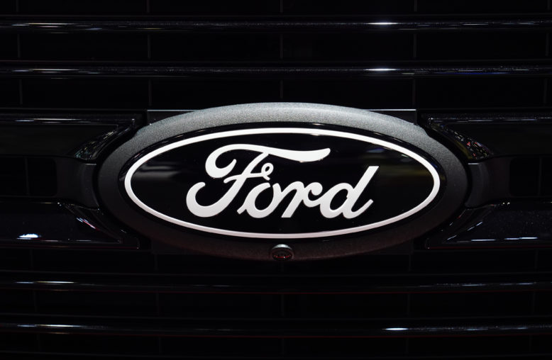 Emblema oval da Ford, preto com letras e bordas brancas, fixado na grade preta de um veículo, com câmera frontal.