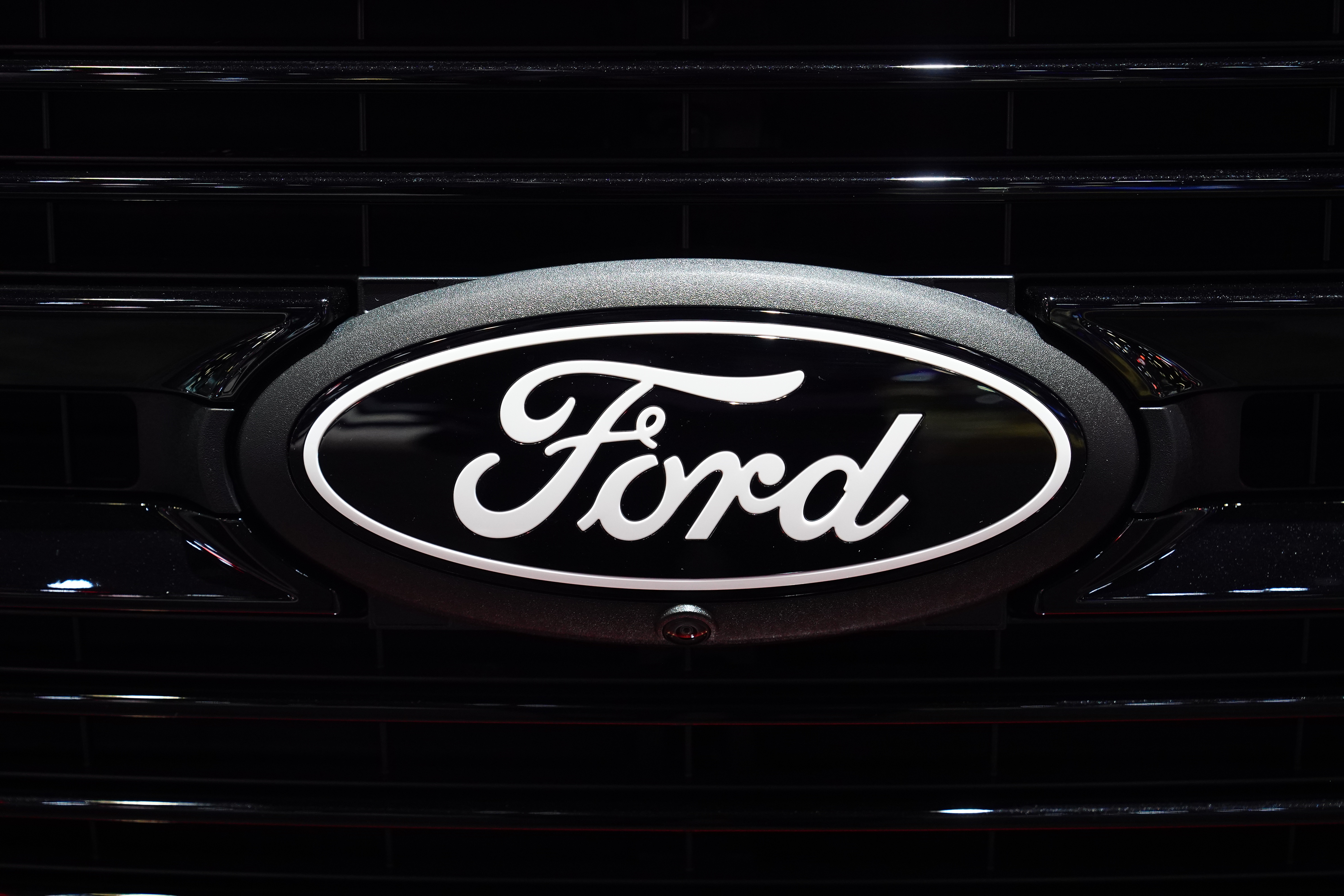 Emblema oval da Ford, preto com letras e bordas brancas, fixado na grade preta de um veículo, com câmera frontal.