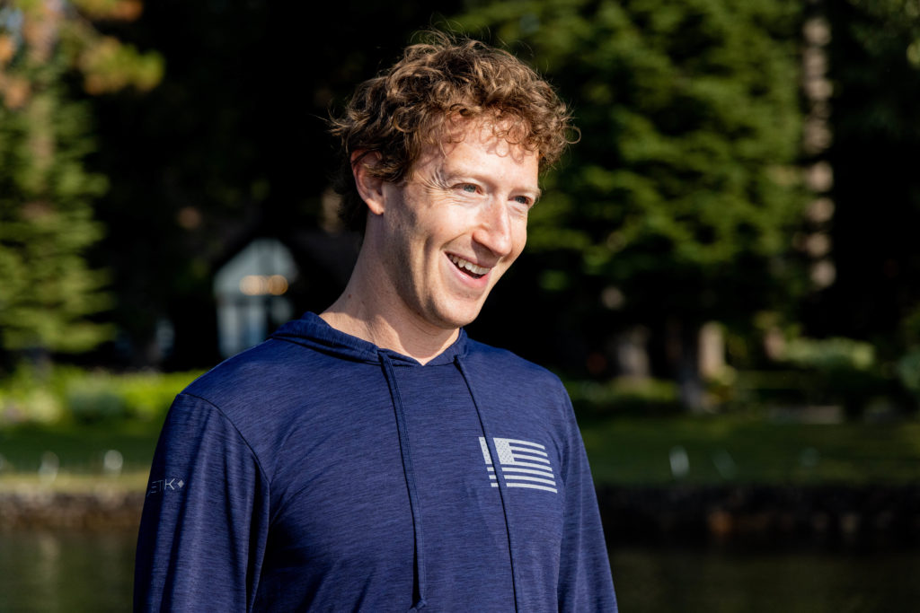 Homem de cabelo encaracolado sorrindo, em moletom azul, com árvores e reflexo de água ao fundo.