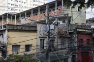 Cena urbana densa com casas empilhadas, muitos fios elétricos em um poste principal e roupas estendidas em varais.