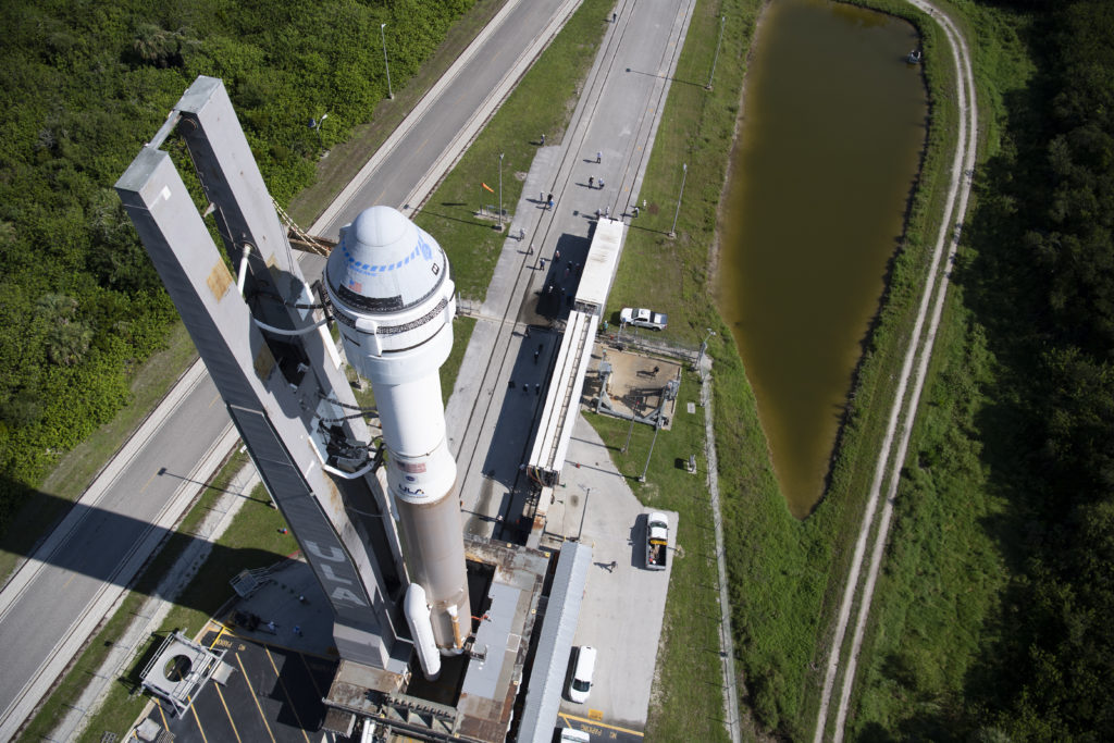 Vista aérea de um foguete Atlas V com Starliner na plataforma de lançamento, cercado por estradas, lago e vegetação.