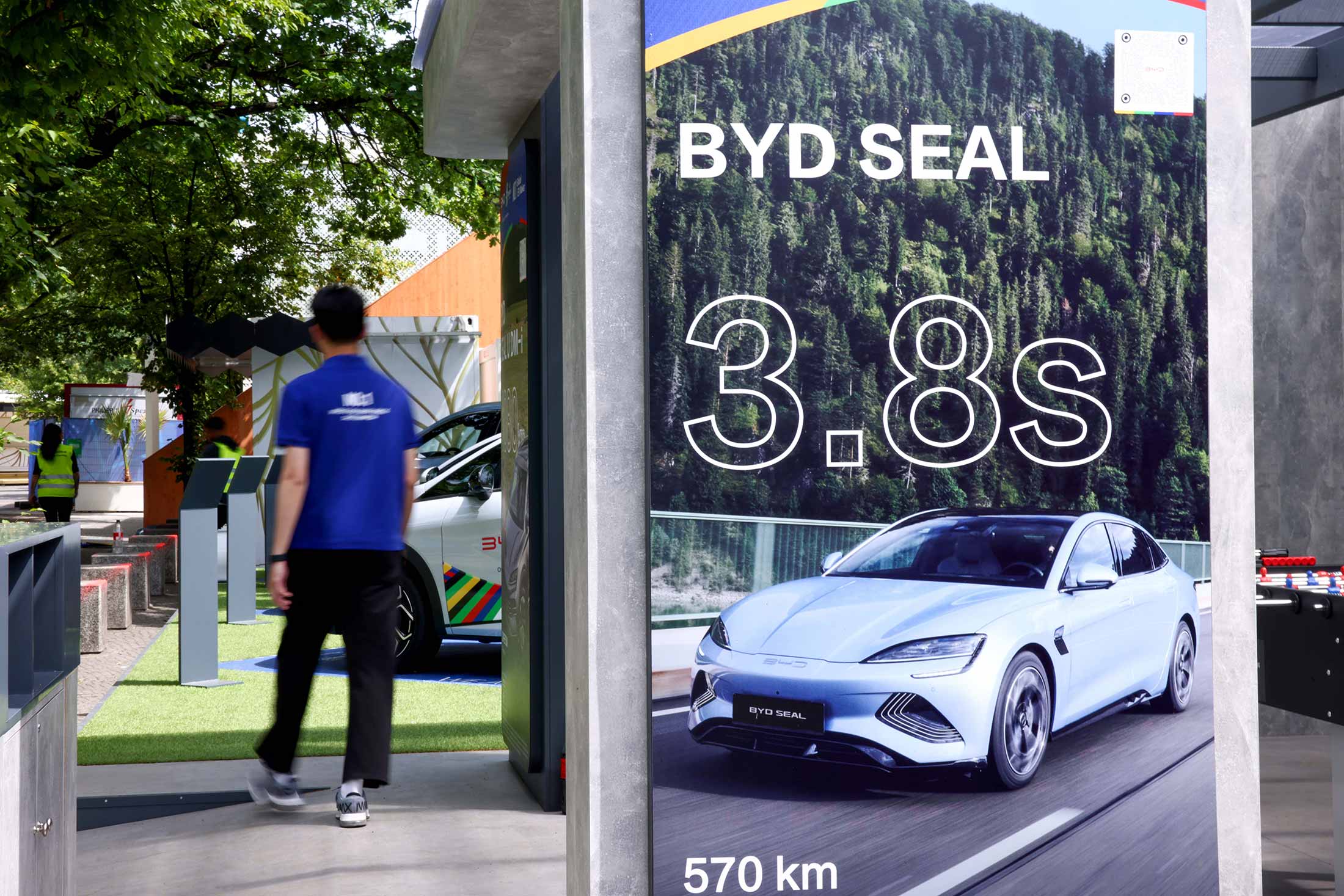 Pôster vertical do BYD SEAL com detalhes de desempenho. Um homem caminha por uma área de exposição de carros.