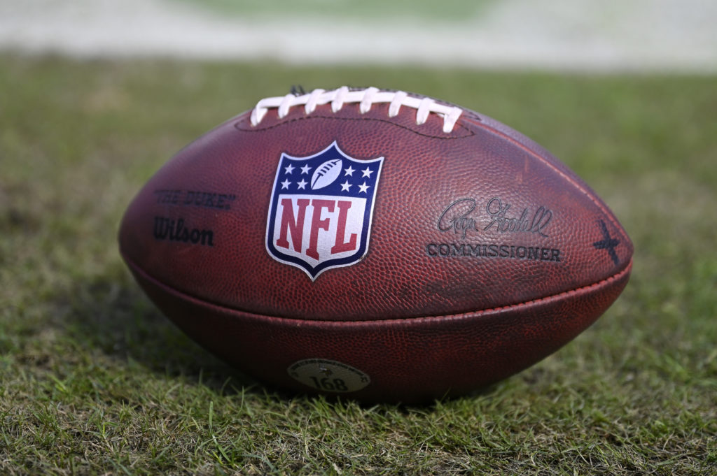 Bola de futebol americano oficial da NFL, em couro marrom e cadarços brancos, pousada na grama.