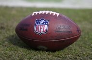 Bola de futebol americano oficial da NFL, em couro marrom e cadarços brancos, pousada na grama.