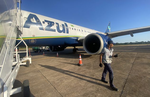 Funcionário da Azul caminha no pátio, próximo a avião da Azul com escada de embarque.