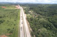 Vista aérea de rodovia movimentada com caminhões e carros, entre campos e floresta densa com casas e morros ao fundo.