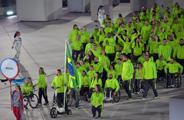 Atletas brasileiros em uniformes verdes vibrantes desfilam, alguns em cadeiras de rodas, com a bandeira do Brasil.