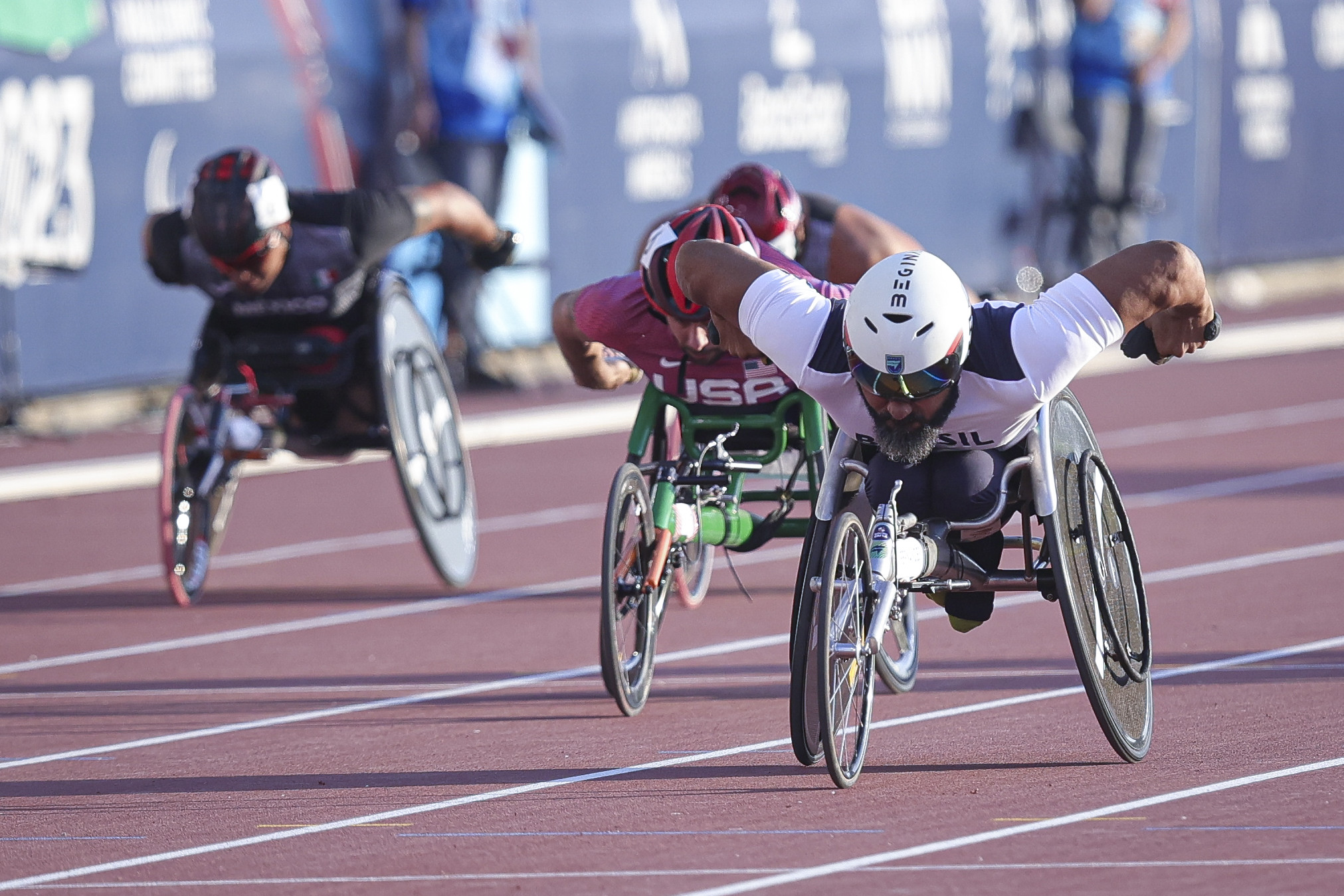 Três atletas em cadeiras de rodas de corrida acelerando em pista de atletismo.