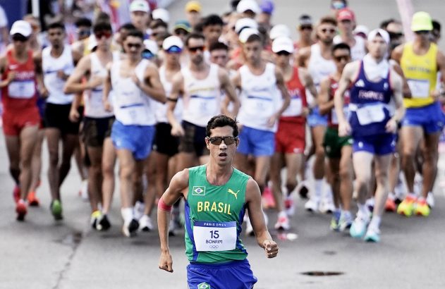Atleta brasileiro de marcha atlética em primeiro plano, com uniforme verde e óculos. Grupo de outros atletas ao fundo.
