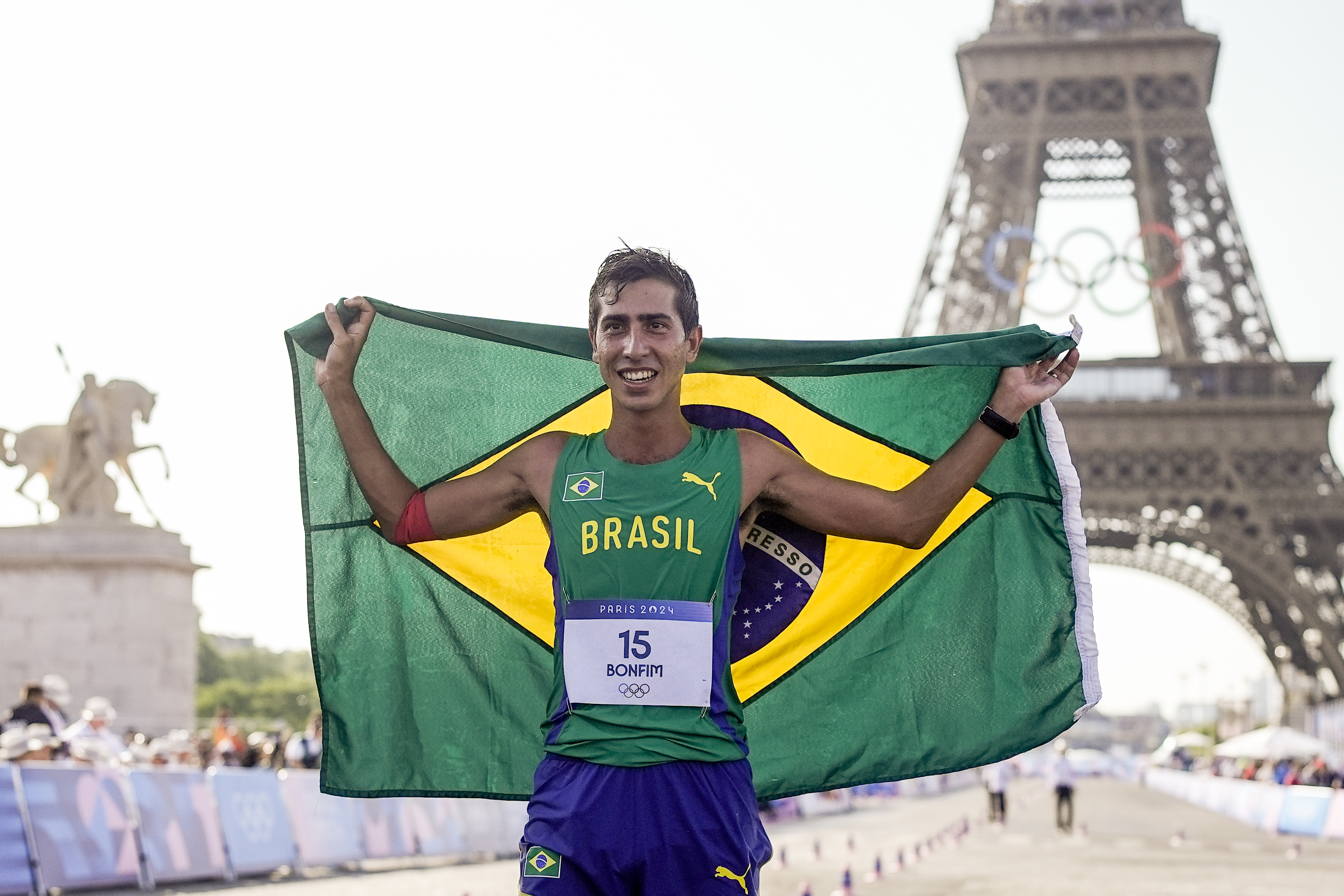 Atleta sorridente segura bandeira do Brasil com Torre Eiffel ao fundo.