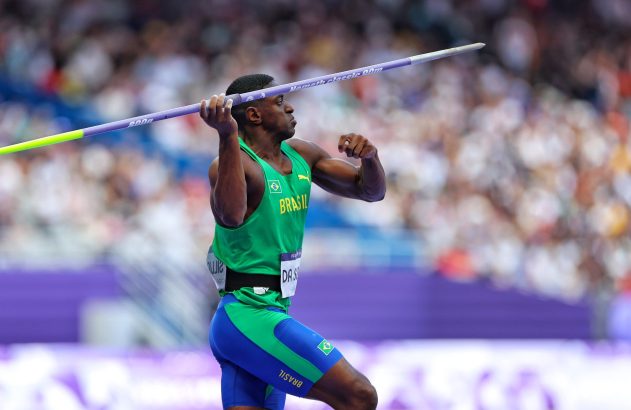 Atleta brasileiro em uniforme verde e azul, preparando lançamento de dardo em estádio.